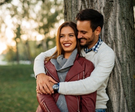Smiling woman and man hugging each other outdoors next to tree