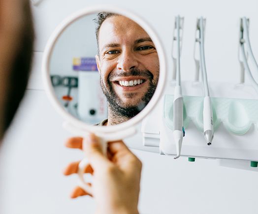 Man smiling at reflection in handheld mirror in dentist's office