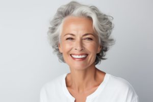 Woman with gray hair in a white shirt smiling at the camera in front of a gray white background